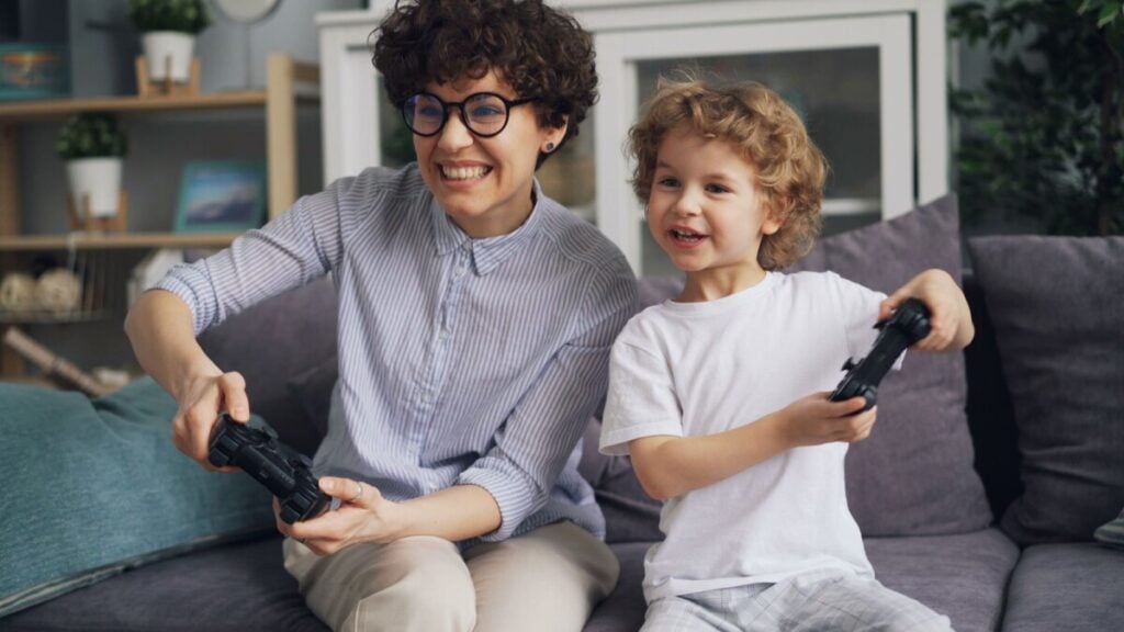 A mother and son smiling and playing video games on a sofa indoors.